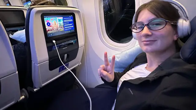 Camila, da Desenrola, faz sinal de paz no assento do avião, com a tela de bordo ao lado, na viagem para Orlando