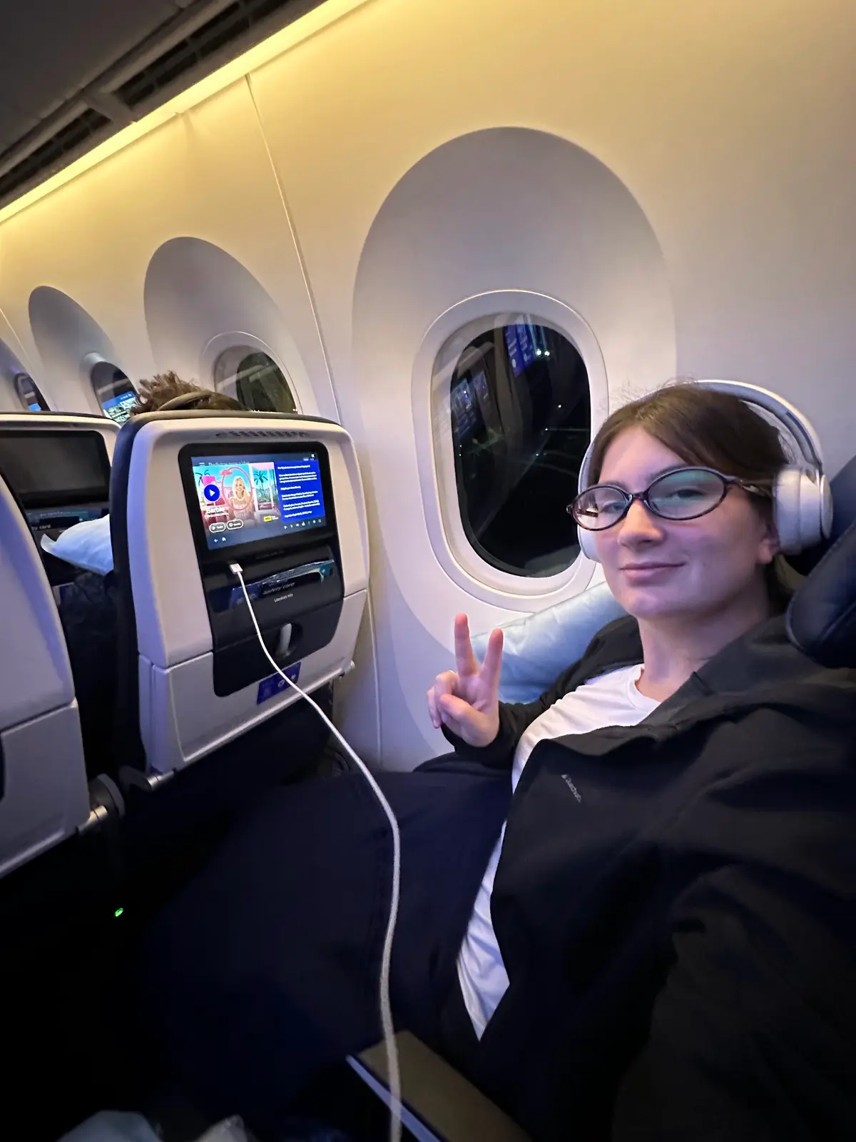 Camila, da Desenrola, no avião com fones de ouvido, fazendo sinal de paz, na viagem para Orlando