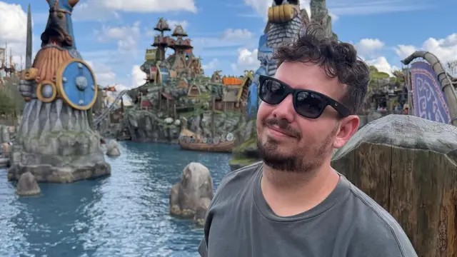 Thomaz sorri na Isle of Berk, no Epic Universe da Universal Orlando, com a lagoa e estátuas vikings ao fundo