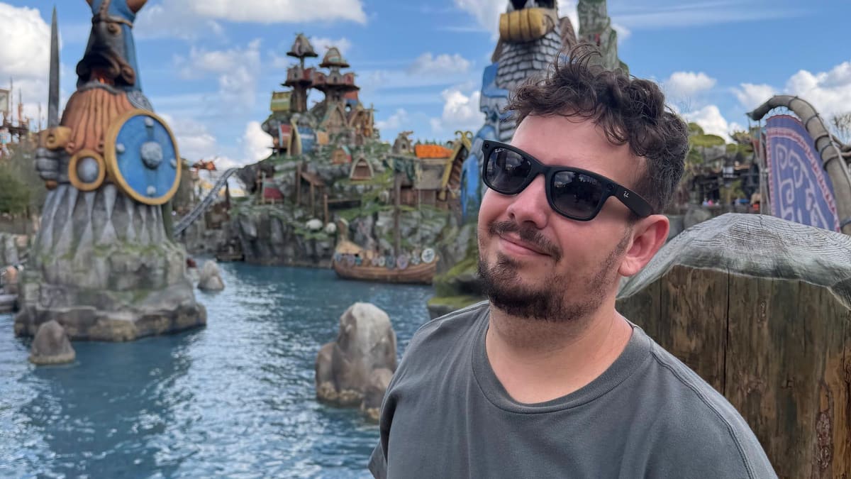 Thomaz sorri na Isle of Berk, no Epic Universe da Universal Orlando, com a lagoa e estátuas vikings ao fundo