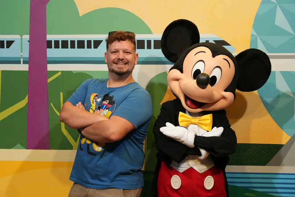 Thomaz de braços cruzados, sorrindo ao lado do Mickey, que imita a pose, diante de um painel com monotrilho e Spaceship Earth estilizada