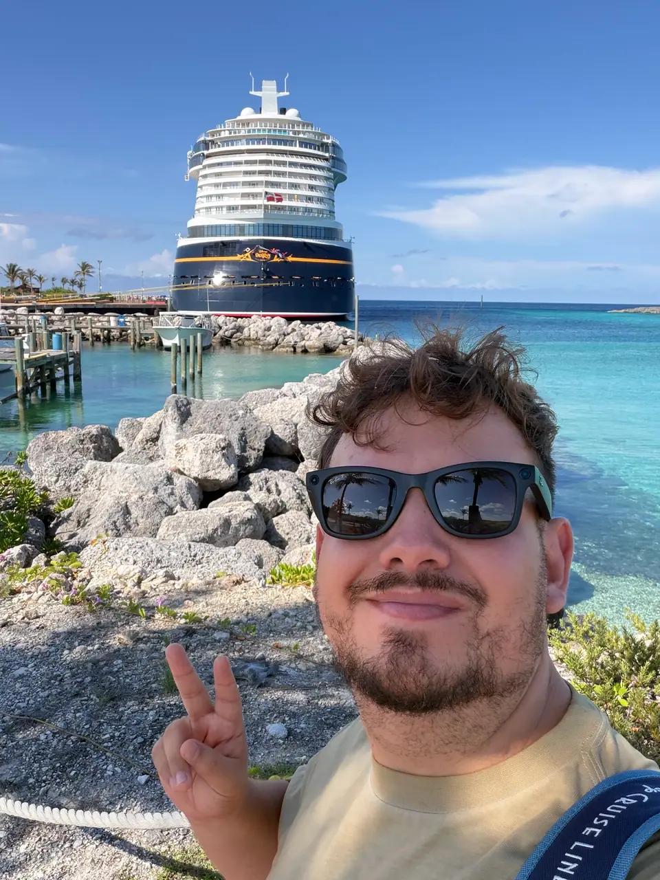 Selfie de Thomaz de óculos escuros fazendo sinal de paz em Castaway Cay, navio Disney Destiny e mar turquesa ao fundo
